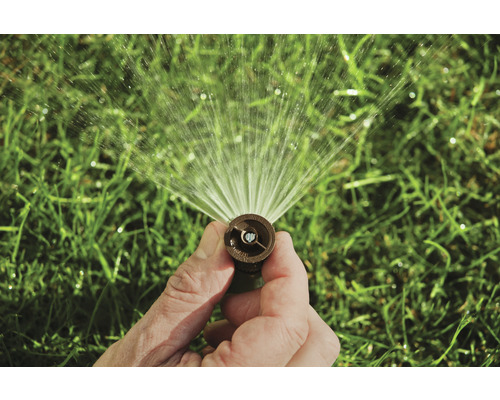 Ruka drží zavlažovač trávníku, který stříká vodu.