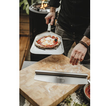 Muž drží pizzu na lopatě na pizzu před prkénkem s kráječem na pizzu