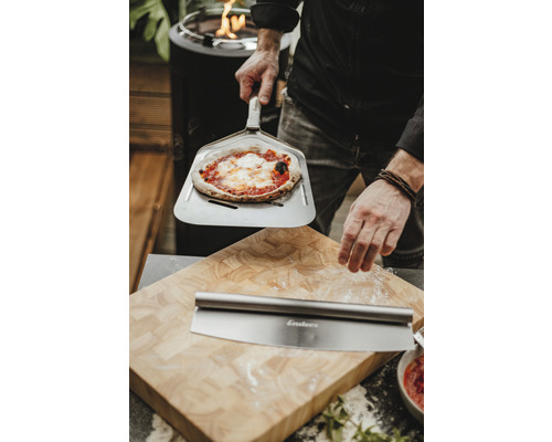 Muž drží pizzu na lopatě na pizzu před prkénkem s kráječem na pizzu