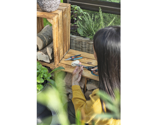 Žena stříhá provázek zahradnickými nůžkami vedle dřevěných beden a rostlin.