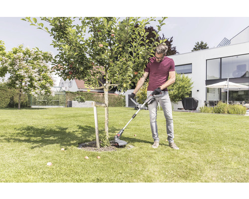Muž zastřihuje trávník strunovou sekačkou kolem stromu na zahradě.