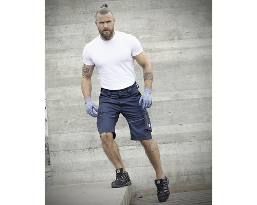 Muž v kraťasech, tričku, rukavicích a bezpečnostní obuvi před betonovou zdí.