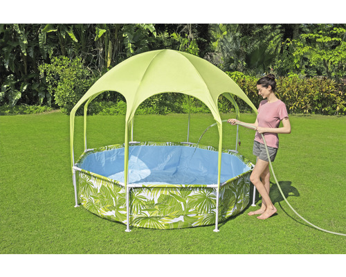 Zahradní scéna s bazénem s rámovou konstrukcí a vzorem listů a sluneční stříškou, zatímco žena napouští bazén zahradní hadicí.