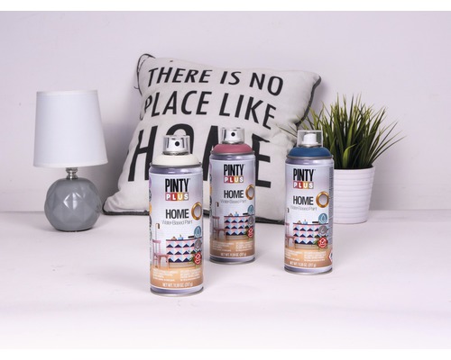 Tři plechovky barvy na vodní bázi Pinty Plus Home před lampou, polštářem a rostlinou