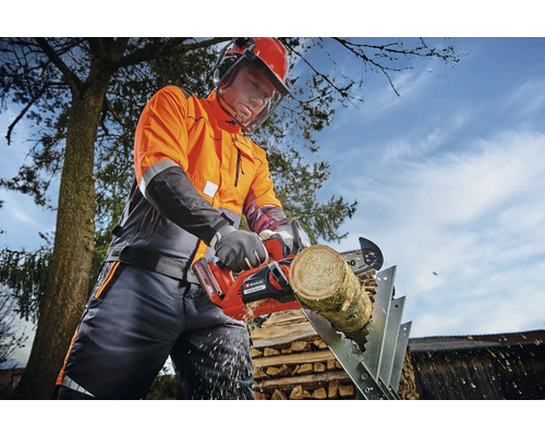 Osoba v ochranném oděvu řeže akumulátorovou motorovou pilou kus dřeva na kozě.