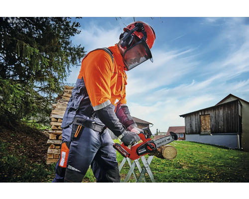 Osoba s ochrannou přilbou a chrániči sluchu pracuje venku s akumulátorovou pilou.