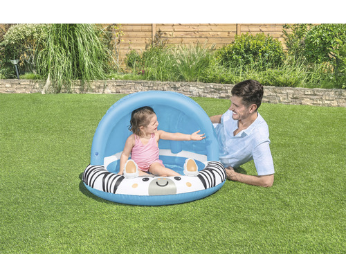 Dítě v bazénku se zebřím vzorem a sluneční ochranou, dospělý leží vedle na trávníku