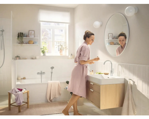 Světlá koupelna s vanou, skříňkou pod umyvadlo a ženou před zrcadlem