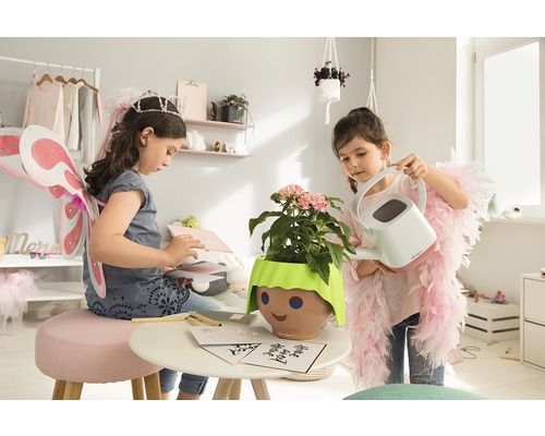 Dvě dívky si hrají v pokoji s rostlinou v květináči s obličejem a konví.