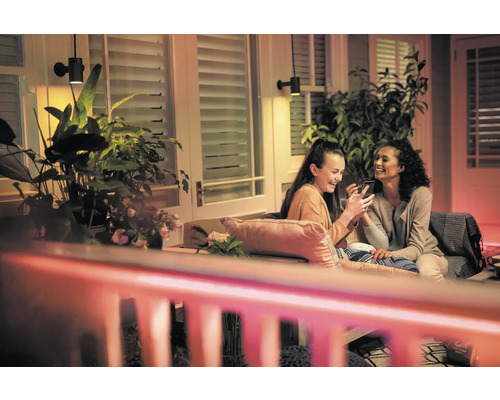 Scéna se dvěma ženami sedícími na terase pod venkovními nástěnnými svítidly, které se dívají na smartphone, obklopené rostlinami.