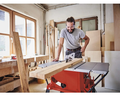 Muž s ochrannými brýlemi řeže dřevěnou desku stolní pilou v dílně.