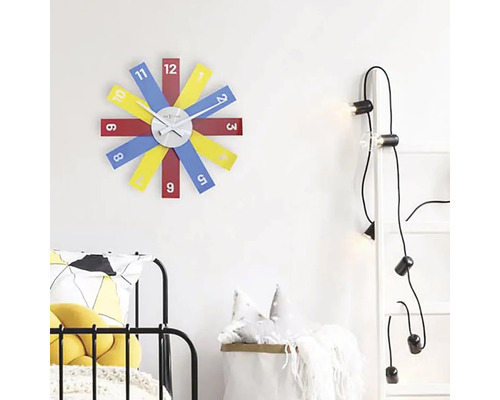 Scéna s nástěnnými hodinami s barevnými ciferníky, žebříkem se žárovkami a postelí.