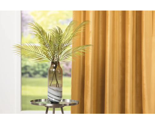 Dekorativní umělá rostlina ve skleněné nádobě na odkládacím stolku před oknem se závěsem.