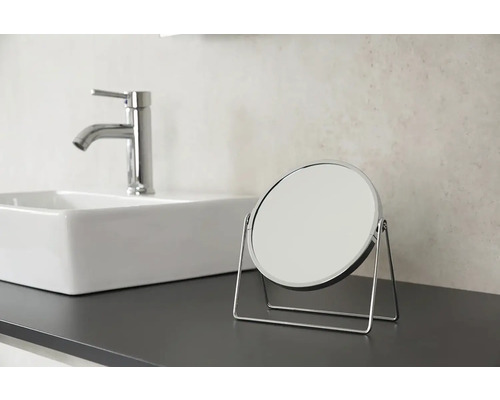 Kulaté kosmetické zrcadlo s chromovaným rámem na desce umyvadla vedle umyvadla a baterie.