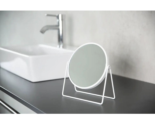 Kulaté kosmetické zrcátko s kovovým rámem na koupelnové poličce s umyvadlem a vodovodní baterií v pozadí.