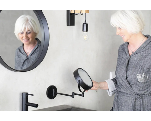 Koupelna s kulatým zrcadlem, černým kohoutkem, lampou a ženou, která se dívá do kosmetického zrcátka.