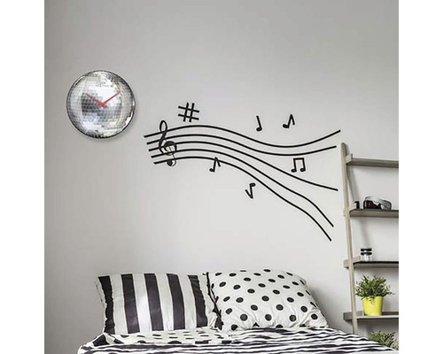 Nástěnná dekorace s zrcadlovými hodinami a nálepkou na zeď s notovými linkami nad postelí.