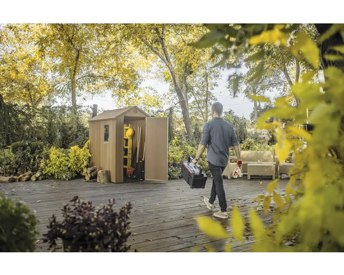 Scéna se zahradním domkem na zahradě a osobou nesoucí bednu s nářadím.