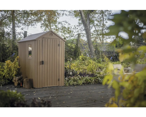 Plastový zahradní domek s oknem a dveřmi na dřevěné terase, obklopený stromy a rostlinami.