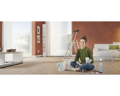 Žena sedí na podlaze v místnosti s kbelíky, žebříkem a renovačními nástroji