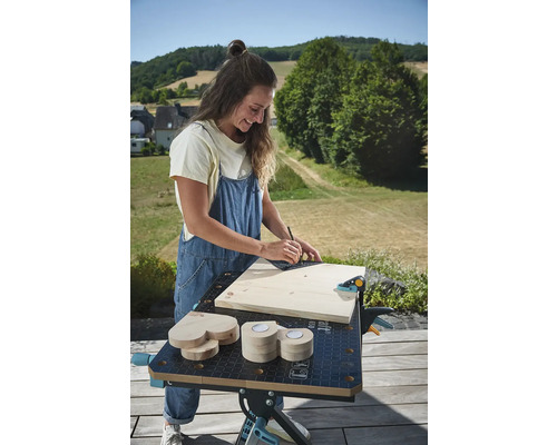 Žena pracuje venku na pracovním stole s dřevem, úhelníkem a tužkou.