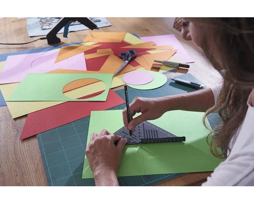 Osoba kreslí úhel a tužku na zelený list papíru, obklopený různobarevným papírem, nůžkami a řezačkou.