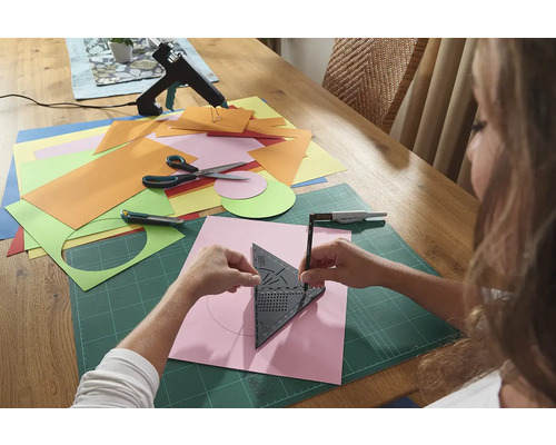 Žena kreslí kruh pomocí trojúhelníku s úhloměrem na kus papíru, obklopeného řemeslnými materiály na stole.