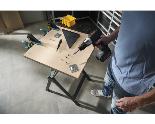 Muž pracuje s akumulátorovým šroubovákem na pracovní lavici se šrouby, úhelníkem a svorkami.