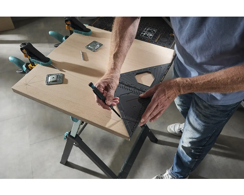 Osoba kreslí úhloměrem a perem čáru na dřevěnou desku, která je připevněna svorkami a kovovými deskami k pracovnímu stolu.