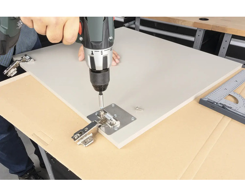 Muž připevňuje závěs pomocí akumulátorového šroubováku k desce.