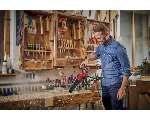 Muž používá akumulátorový foukač listí k odfouknutí třísek z pracovního stolu. V pozadí nářadí ve skříni.