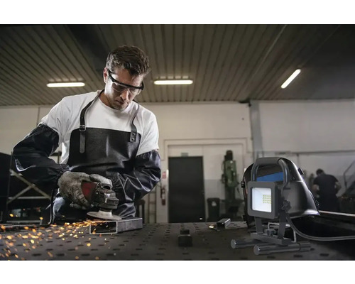 Muž s ochrannými brýlemi, zástěrou a rukavicemi brousí kov pomocí flexy, zatímco v popředí je staveništní reflektor.