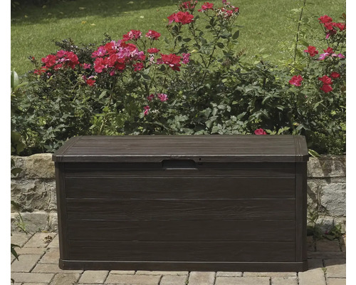 Tmavě hnědý zahradní box před růžovými keři a kamennou zdí.