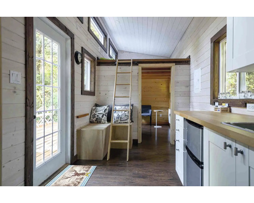 Interiér malého domu s dřevěnými stěnami, žebříkem do zvýšeného patra a kuchyňským koutem.