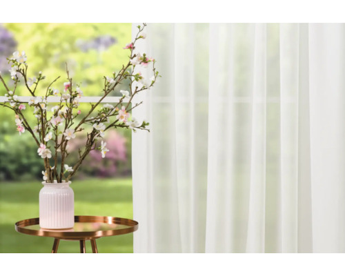 Dekorativní větve s květy ve váze na odkládacím stolku před oknem se závěsem.