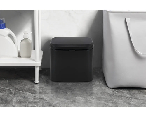 Černý odpadkový koš v koupelně vedle regálu a koše na prádlo.