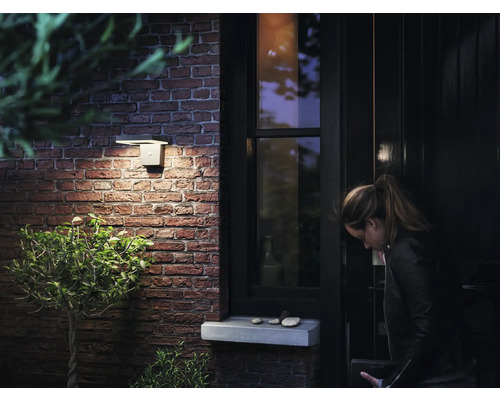 Žena odemyká vchodové dveře, vedle kterých je na cihlové zdi připevněno venkovní svítidlo