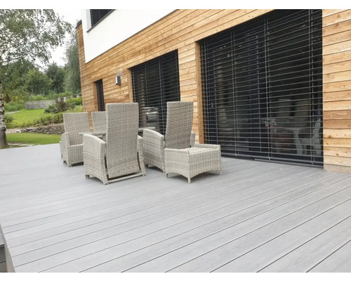 Terasa s šedou dřevěnou podlahou a ratanovým nábytkem před domem s dřevěnou fasádou.