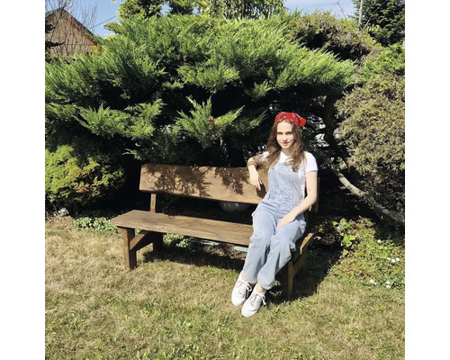 Žena sedí na dřevěné zahradní lavičce před zeleným keřem.