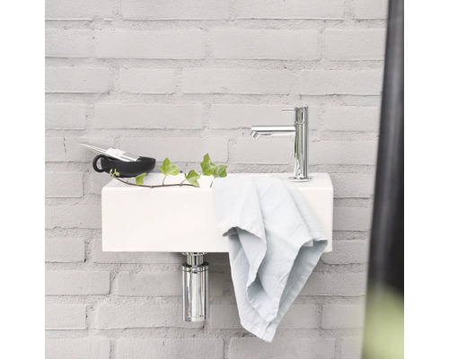 Umyvadlo s baterií, ručníkem, šálkem a rostlinou na světlém cihlovém pozadí.