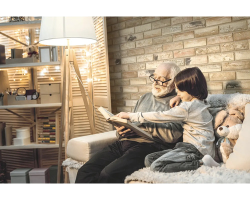 Dědeček čte se svým vnukem knihu v útulném pokoji s policemi, stojací lampou a cihlovou zdí.