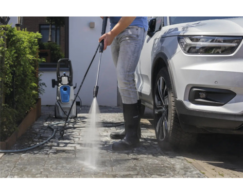 Osoba používá vysokotlaký čistič k čištění příjezdové cesty vedle auta.