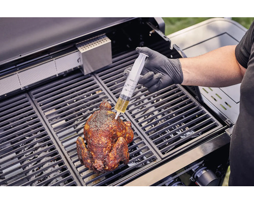 Osoba injekční stříkačkou vstřikuje tekutinu do grilovaného kuřete na grilu.