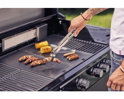 Osoba griluje klobásy, slaninu a kukuřičné klasy na grilu.