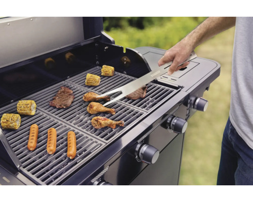 Gril s grilovanými potravinami, jako jsou steaky, klobásy, kuře a kukuřičné klasy, se obrací grilovacími kleštěmi.