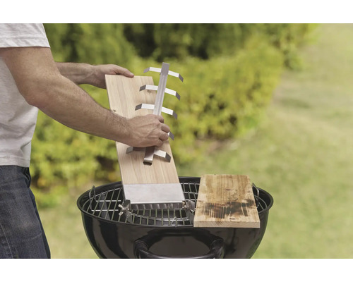 Scéna s mužem držícím grilovací desku s držákem nad grilem na dřevěné uhlí, venku.