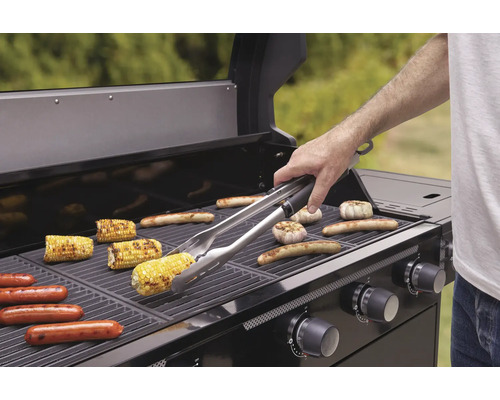 Gril s kukuřičnými klasy, klobásami a česnekem na grilovacím roštu a osoba s grilovacími kleštěmi.