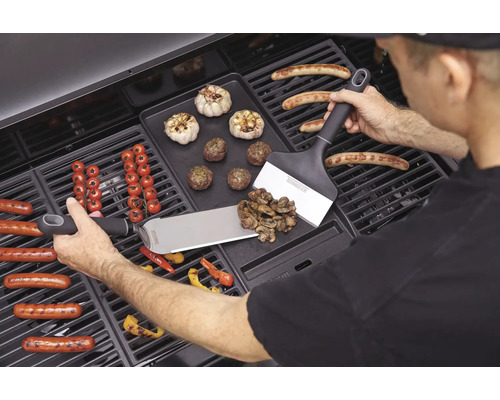 Muž griluje klobásy, zeleninu a česnek na grilu s obracečkami.