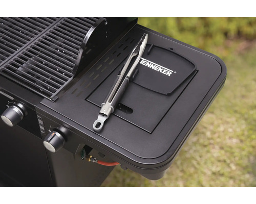 Detailní pohled na černý plynový gril s grilovacími kleštěmi na boční polici a logem Tenneker.