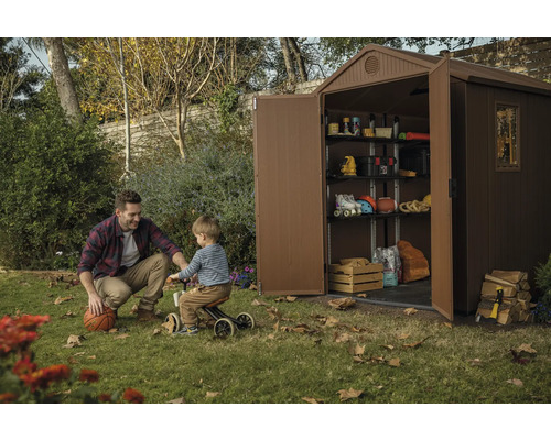 Scéna s plastovým zahradním domkem s policemi, který je plný různých věcí, zatímco si muž a dítě hrají na trávníku.